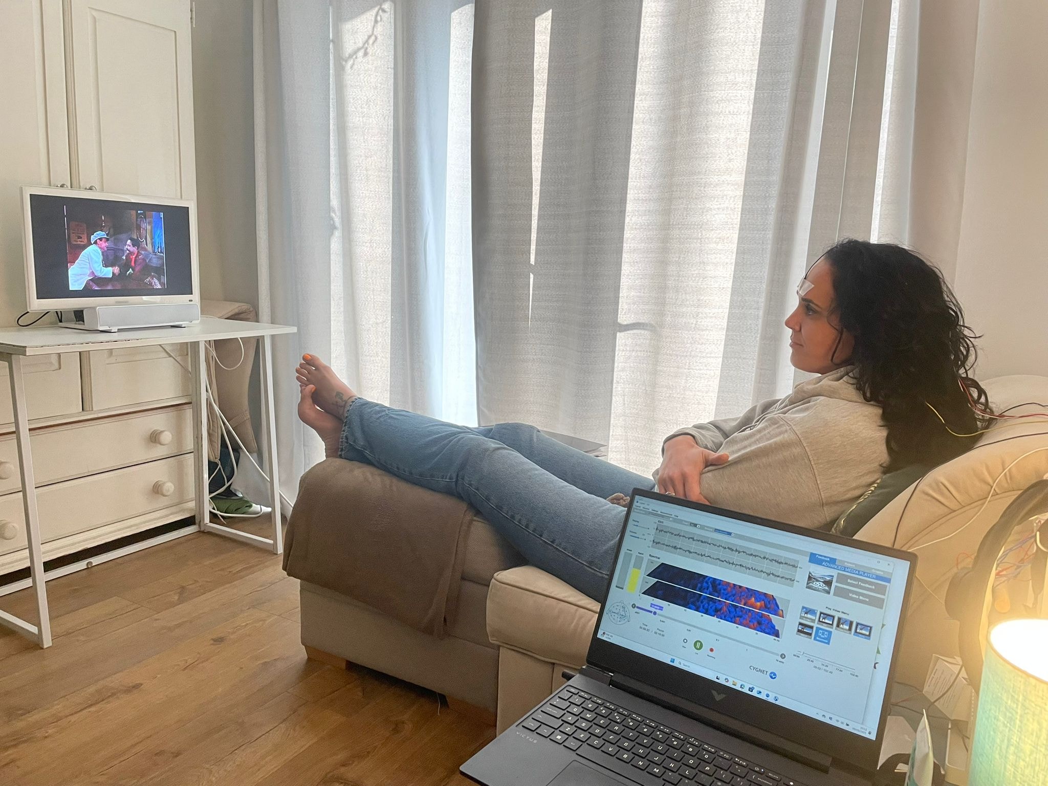 A client relaxing in a chair watching TV during a neurofeedback therapy session, with sensors on her head and brainwave monitoring software visible on a laptop